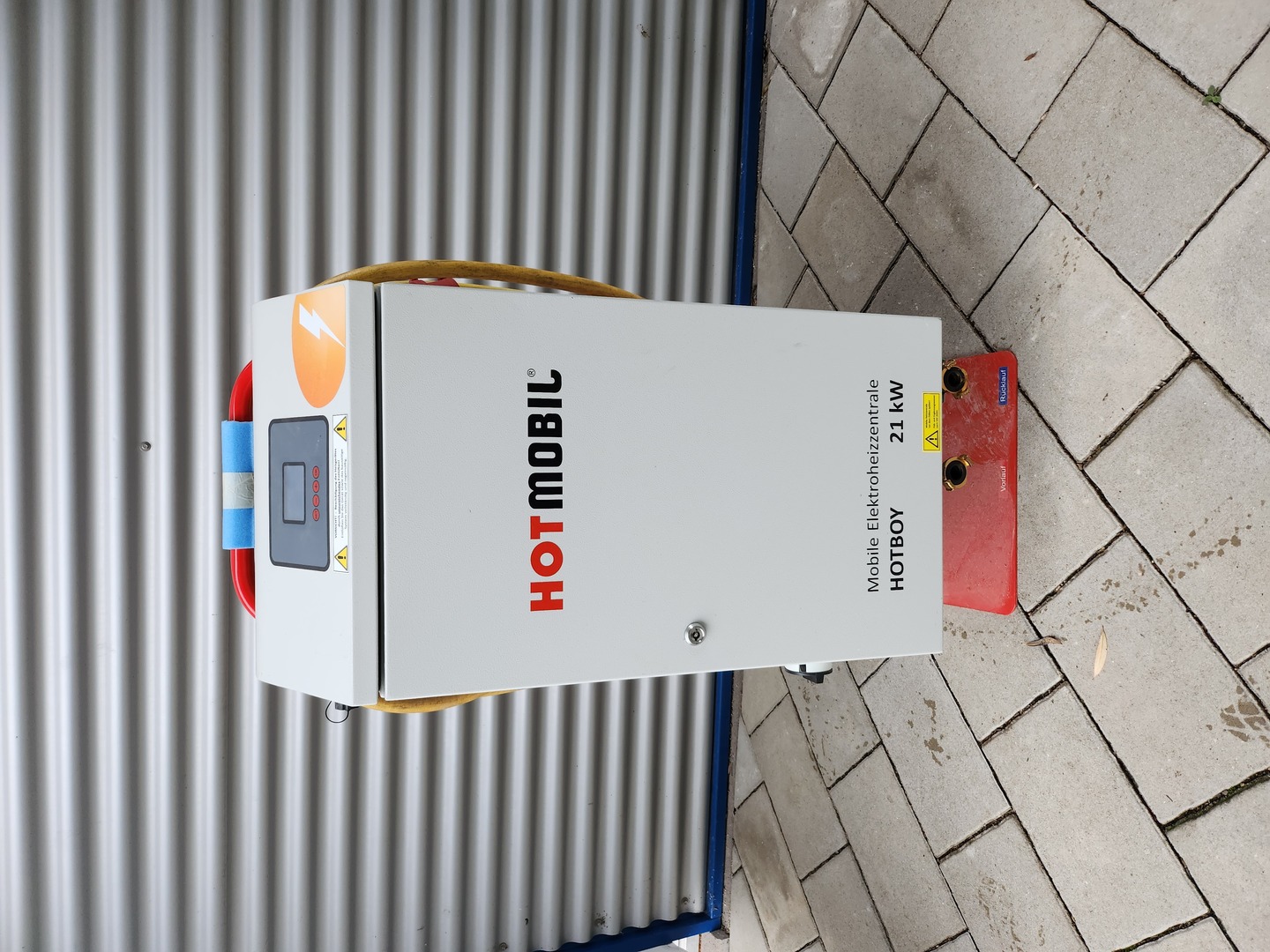 Ein weißer Stromverteiler mit der Aufschrift "HOTMOBIL Mobile Elektrohilfzentrale HOTBOY 21 kW" steht auf einem gepflasterten Boden neben einem Metallcontainer.