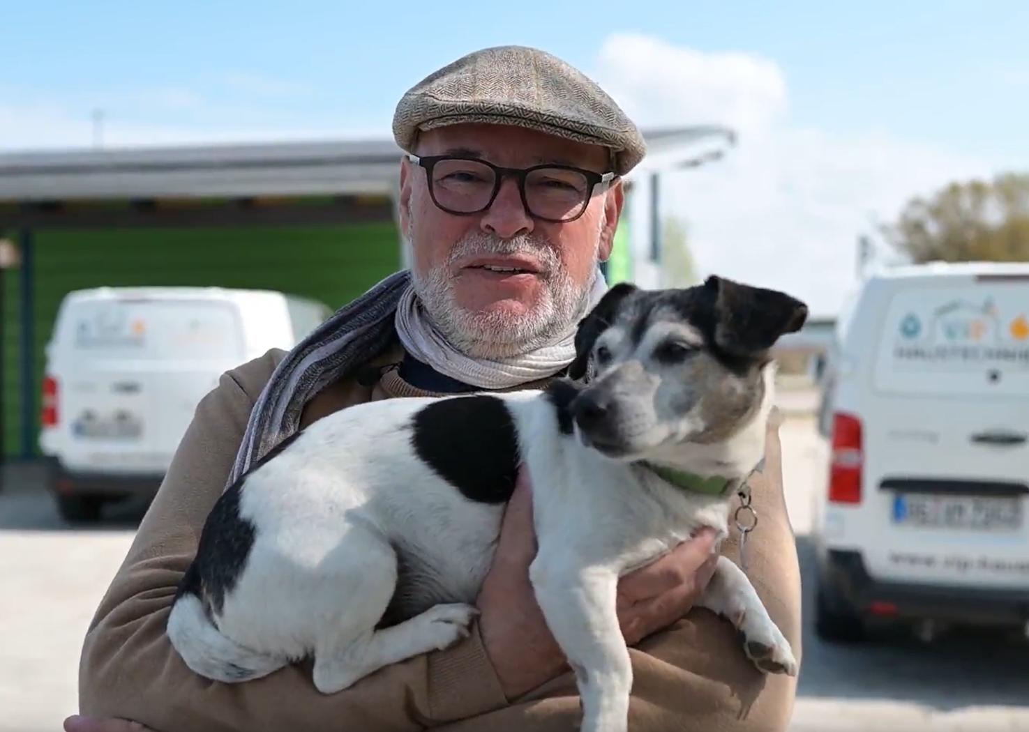 Ein Mann mit Brille und Mütze hält einen kleinen Hund mit schwarz-weißem Fell im Arm.