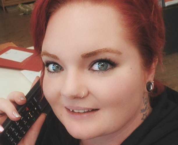 Eine Person mit roten Haaren, Piercings und einem Tattoo am Hals hält ein Telefon und lächelt.